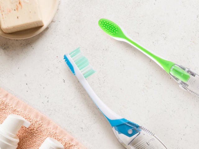 Two GUM TARVEL toothbrushes in blue and green colours, shown from the side and back, placed in a natural environment.